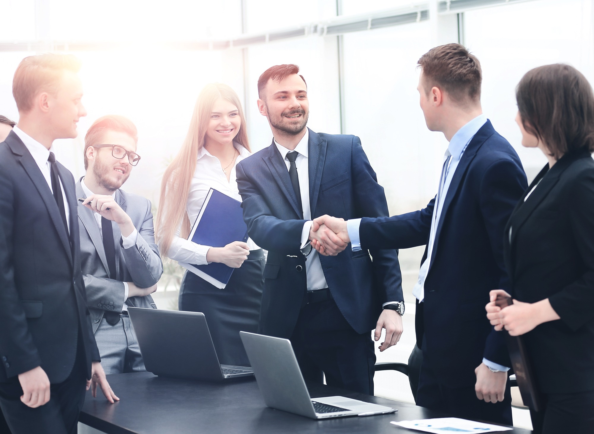 Business professionals engage in a handshake during a meeting, showcasing collaboration and agreement. Laptops and a notebook indicate a formal setting.