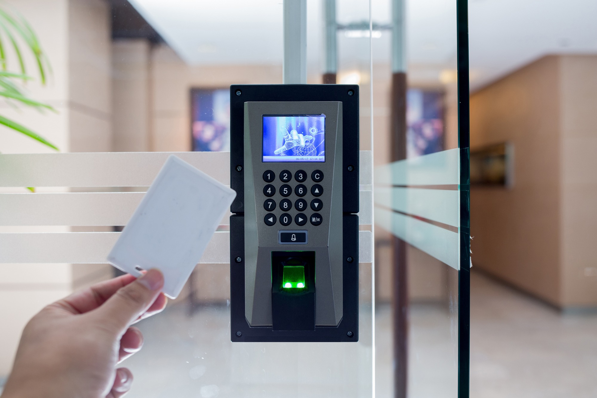 Person holding a card near a biometric access control panel, featuring a keypad and digital display, used for secure entry.